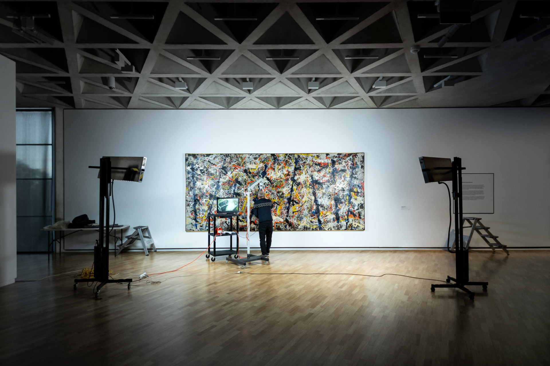 The gallery is dark. There are two large lights on Blue poles. Senior Painting Conservator David Wise is inspecting the painting.